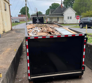 A dumpster filled with construction waste including wood and metal scraps parked on a street beside a building.